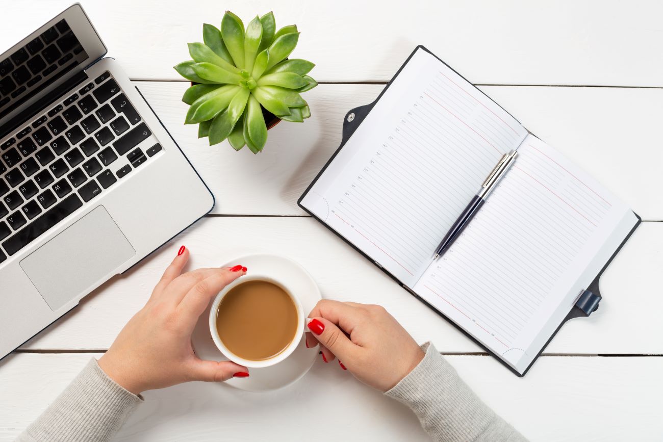 woman-with-red-nails-holding-cup-coffee-near-modern-laptop-plant-pot-notebook-with-pen